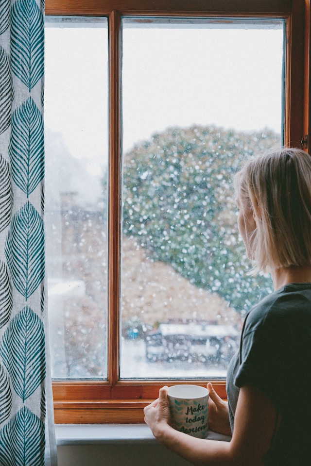 Woman looks out window that has raindrops on it. She has short blond hair. She is holding a mug that says "make today awesome"